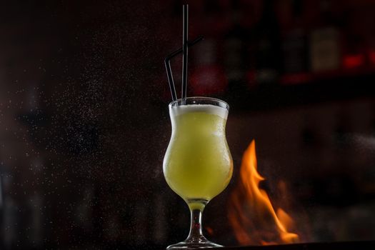Bartender sprinkles on illuminated glass with bright green cold cocktail on bar counter and makes fire flame over it.
