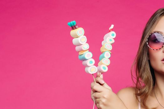 Crop of beautiful and seductive girl wearing in pink sunglasses with makeup like doll, posing at studio with marshmallow candy on stick. Pretty woman in violet sunglasses holding candies. Fashion.