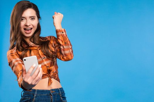 Front view of lucky girl in checkered shirt and jeans looking at camera and taking selfie on blue isolated background. Young woman laughing in studio. Concept of happiness and technology.