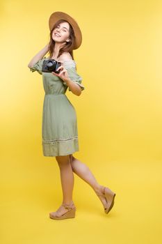 Front view of bright young girl wearing straw hat and glasses posing on yellow isolated background in studio. Female tourist keeping camera, looking at camera and smiling. Concept of summer.