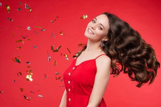 Portrait of adorable smiling young woman long curly hair posing isolated at red studio background. Beautiful happy female enjoying relaxing surrounded by shining colorful confetti looking at camera