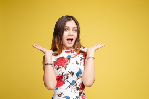 Front view of surprised young girl in bright dress looking at camera on yellow isolated background in studio. Funny amazed female with open eyes shouting. Concept of shock and happiness.