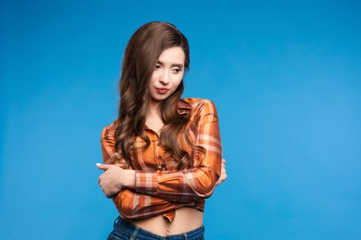 Front view of shy beautiful woman in orange checkered shirt and jeans posing on blue isolated background in studio. Young model with folded arms looking down. Concept of outfit and lifestyle.