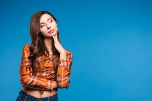 Front view of beautiful woman wearing checkered shirt looking aside and thinking on blue isolated background. Young long haired girl keeping fingers near chin and dreaming. Concept of workday.