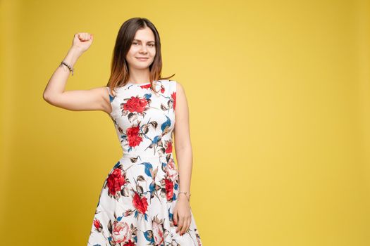 Studio portrait of fashionable brunette lady in white dress with flowers and beige heels posing with bent leg. Smiling at camera. holding skirt. Isolate on yellow background.