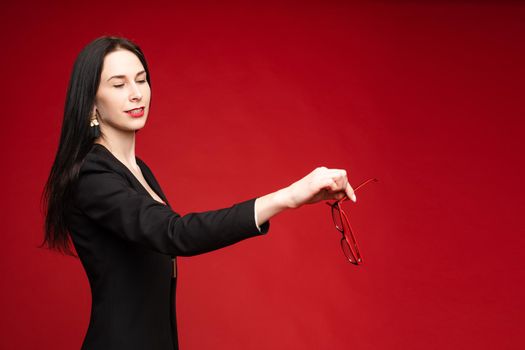 Studio portrait of adult brunette model with red lips wearing black jacket throwing her old glasses away. Eye correction concept. Isolate on red.