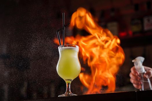 Bartender sprinkles on illuminated glass with bright green cold cocktail on bar counter and makes fire flame over it.