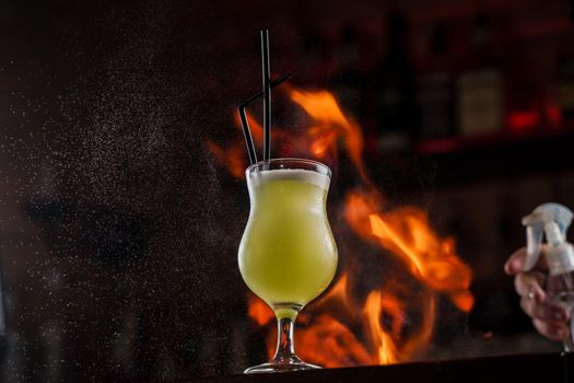 Bartender sprinkles on illuminated glass with bright green cold cocktail on bar counter and makes fire flame over it.