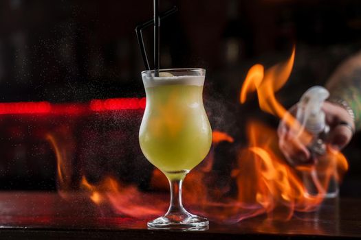 Bartender sprinkles on illuminated glass with bright green cold cocktail on bar counter and makes fire flame over it.