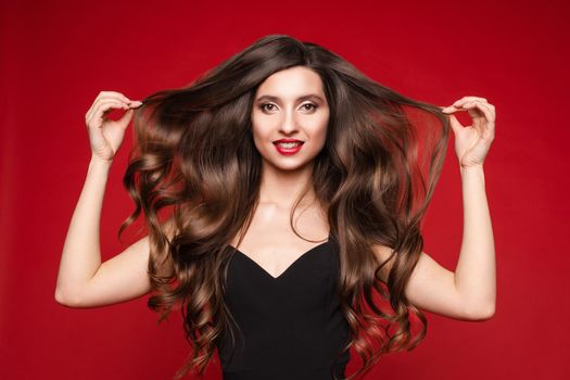 Studio portrait of stunning young caucasian woman with gorgeous long brunette hair in black with red lips. Isolate on red. She is showing beautiful, smooth and healthy hair.