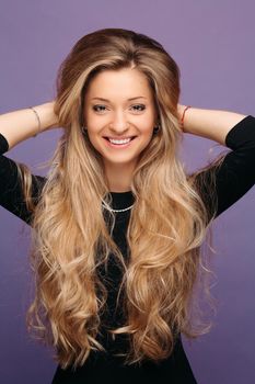 Portrait of young blonde model with long volumed hair and perfect makeup after beauty salon posing and smiling at camera. Pretty girl touching hair, posing on violet studio background.