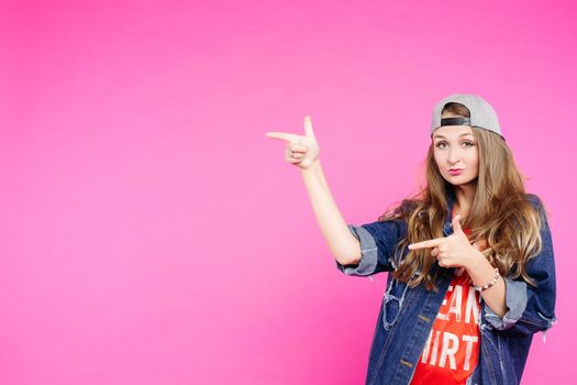 Young trendy teen girl, pointing at side, showing by fingers and looking at camera. Funky and swag blondie in cap with long hair making duck face. Concept of teen fashion.