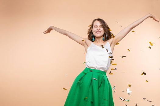 Happy funny woman rising up hand surrounded by falling shining multicolored confetti isolated at light studio background. Adorable smiling female enjoying fun having positive emotion medium long shot
