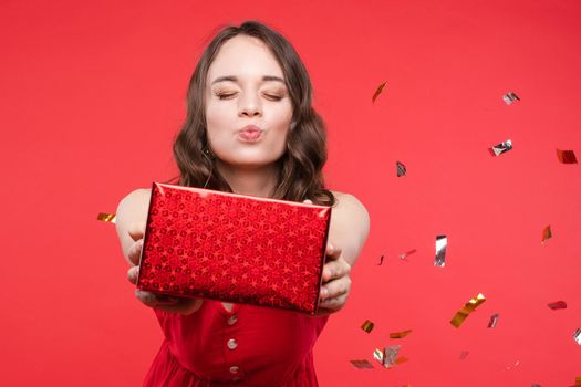 Pleasant young woman taking gift box kissing with closed eyes isolated at red studio background. Happy beautiful young brunette stylish woman posing surrounded by colorful shining confetti medium shot