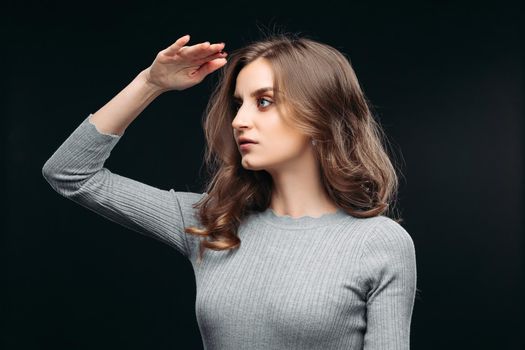 Waist up portrait of waiting attractive woman in nice gray sweater. Lady with long curly hair and delicate makeup is posing in studio. Isolated on dark background