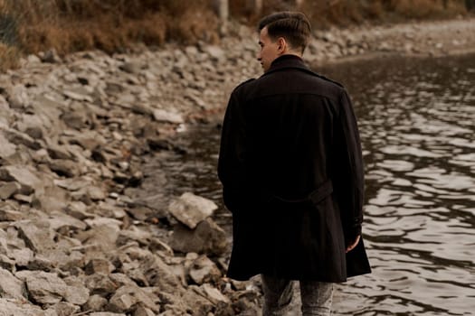 Confident male model weared black fashion coat. Handsome man walks near lake