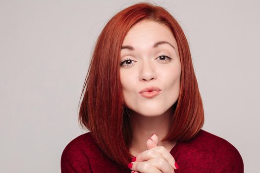 Young pretty lady with red hair in bordo blouse holding eyebrows up and sending kiss. Close up of beautiful woman with red nails holding hadns together near neck. Concept of love to yourself.