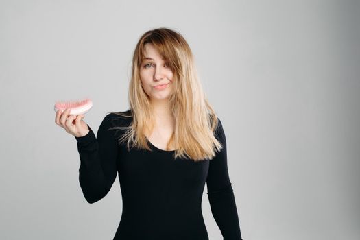 Close up portrait of young confused girl with messy blonde hair. Pretty woman with green eyes, wearing in black sweater, holding hairbrush in hand and looking at camera against white background.