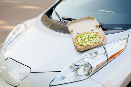 Pizza closeup on hood of the eco car. Safe delivery of pizza with green salad, tomatoes, cheese. Ecological delivering by electric car.