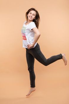 Full length isolate portrait of positive brunette young girl in jeans and t-shirt and sneakers jumping over yellow background. She is smiling at camera.