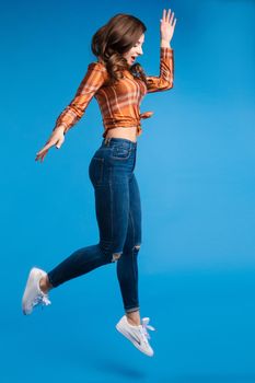 Cool young and beautiful brunette girl in checked shirt tied in front and slim jeans with sneakers jumping in mid-air. She is active and energetic. Isolate on blue.