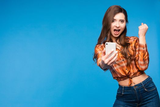 Front view of lucky girl in checkered shirt and jeans looking at camera and taking selfie on blue isolated background. Young woman laughing in studio. Concept of happiness and technology.