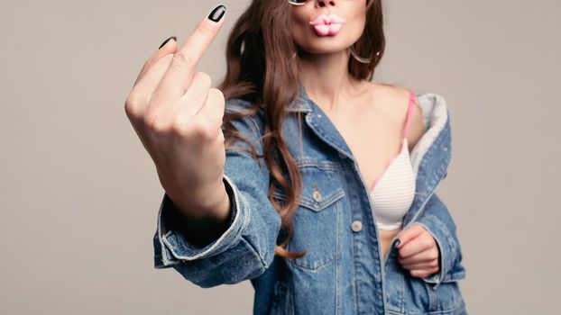 Offensive gesture showing at camera. Stylish young brunette in mirrored sunglasses showing middle finger with black nail. She is wearing denim jacket over a bra. Isolate on grey. Studio shot.