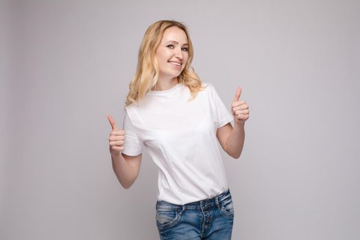 Front view of positive blonde wearing white shirt and jeans looking at camera and showing thumbs up. Young woman posing and laughing on grey isolated background. Concept of happiness.