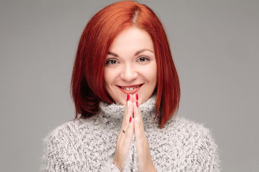 Adorable and beautiful young woman with smooth skin and straight ginger hair posing. Beautiful model looking at camera and smiling. Girl wearing in grey sweater and putting hands together.