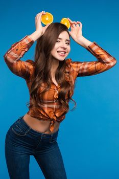 Young girl in casual clothes hiding her face behind fruits. Brunette lady in checkered shirt and jeans holding halves of oranges as sunglasses. Beatiful cute woman with opened belly happily smiling.