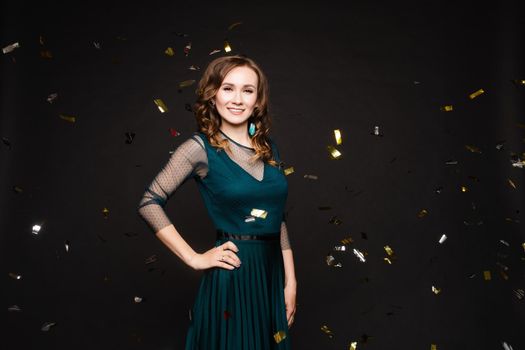 Happy young girl looking surprisingly while golden confetti falling. Beautiful woman in emerald elegant long dress smiling and dancing among decorations. Gorgeous lady celebrating birthday on party.