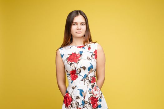 Studio portrait of fashionable brunette lady in white dress with flowers and beige heels posing with bent leg. Smiling at camera. holding skirt. Isolate on yellow background.