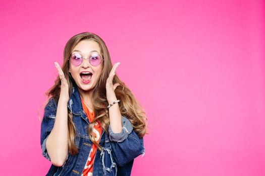 Stylish swag girl wearing jeans jacket with sunglasses, shocked excited screaming with hands to cheeks and open mouth. Pretty and young teenage in pink glasses screaming at studio.