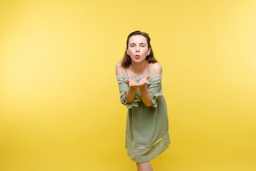 Studio portrait of pretty young Caucasian woman in plain pale green dress blowing an air kiss to camera holding hands together. She is isolated on plain yellow background.