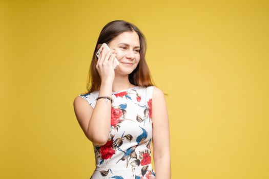 Front view of stylish young girl in bright light dress talking by phone on yellow isolated background in studio. Lovely female chattering and smiling. Concept of communication and happiness.