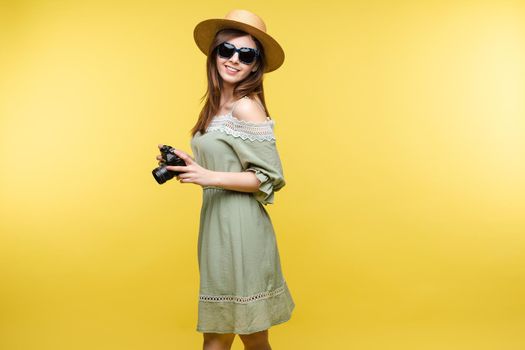 Front view of bright young girl wearing straw hat and glasses posing on yellow isolated background in studio. Female tourist keeping camera, looking at camera and smiling. Concept of summer.