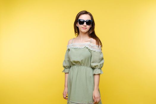 Studio portrait of attractive happy and jovial Caucasian brunette woman in plain pistachio dress and trendy black sunglasses smiling at camera against yellow background.