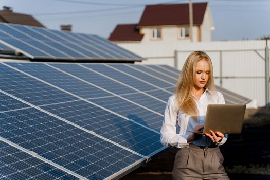 Investor woman stand with laptop near blue solar panels row on the ground. Free electricity for home. Sustainability of planet. Green energy.