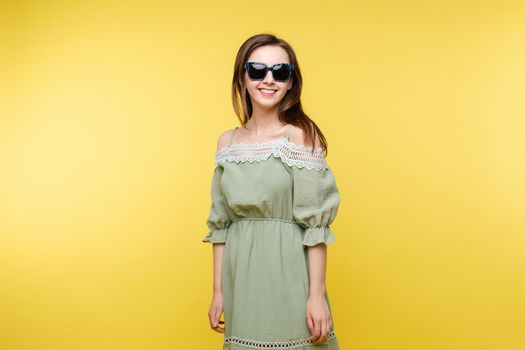 Studio portrait of attractive happy and jovial Caucasian brunette woman in plain pistachio dress and trendy black sunglasses smiling at camera against yellow background.