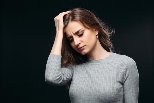 Waist up portrait of sad and worried female in gray sweater holding hand to head. Wailful lovely girl with nice makeup posing at studio. Isolated on dark background