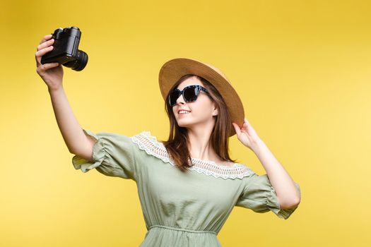 Studio portrait of fashionable young brunette girl in summer dress and stylish hat and sunglasses posing while taking self-portrait via film camera. Isolate on yellow.