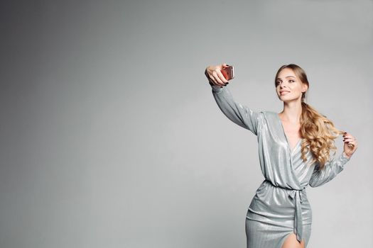 Portrait of a blonde woman dressed in a stylish dress, with make-up and a hairdress after a beauty salon, makes selfies for social networks. A gorgeous smiling girl with twisted hair posing in a gray studio.