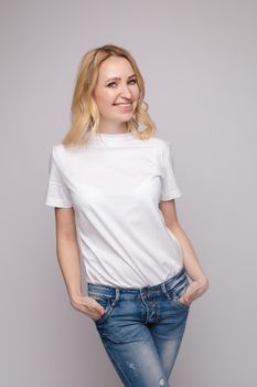 View from front of beautiful slim woman wearing white shirt and jeans standing steady on frey isolated background. Young blonde looking at camera, smiling and posing in studio.