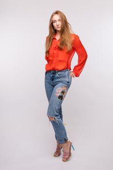 Side view of young pretty girl wearing red blouse and jeans posing on grey isolated background. Longhaired female model standing and looking at camera in studio. Concept of full length.