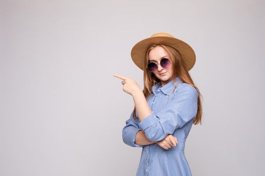 Studio portrait of stylish woman with redhair in sunglasses, hat and checked shirt dress indicating blank space in the air with her index finger.
