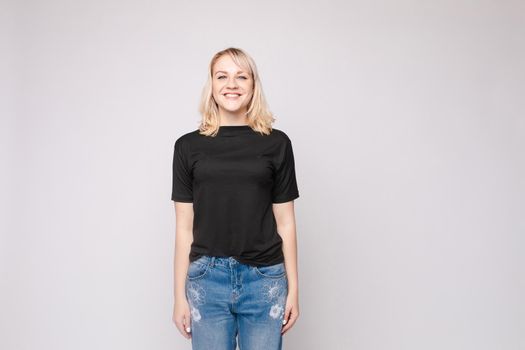 View from front of beautiful slim woman wearing black shirt and jeans standing steady on frey isolated background. Young blonde looking at camera, smiling and posing in studio.