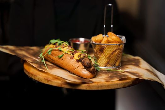 Waiter carries the order to the visitor. Barbecue grilled hot dog with yellow mustard and ketchup on wooden background. Hot dog sandwich with potato fries and sauces.