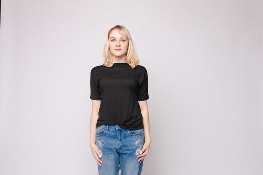 View from front of beautiful slim woman wearing black shirt and jeans standing steady on frey isolated background. Young blonde looking at camera, smiling and posing in studio.