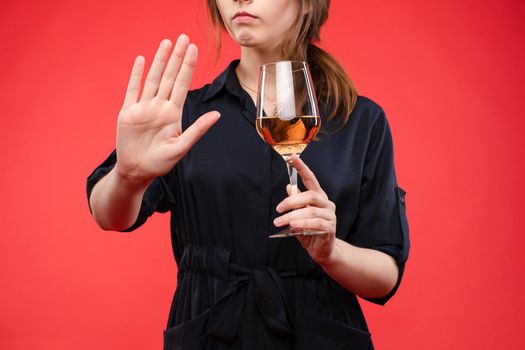 Cropped photo of unrecognizable woman in black dress with a glass of wine showing stop gesture with her hand palm. Isolate over red background.