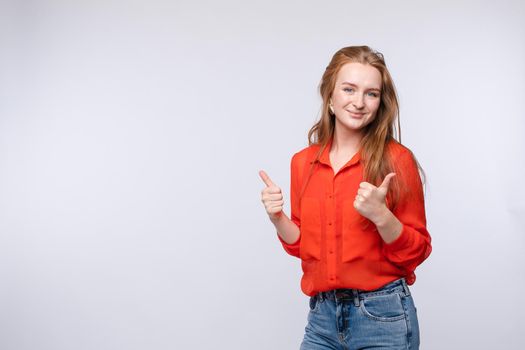 Front view of positive female in red shirt and jeans looking at camera and showing thumbs up on grey isolated background. Longhaired girl gesturing and smiling. Concept of approval and agreement.